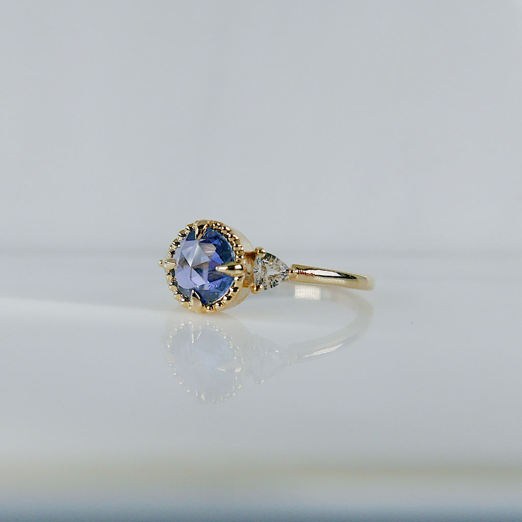 Angled profile photo of a beaded yellow gold ring with compass setting and blue rosecut stone with a clear trillion side stone
